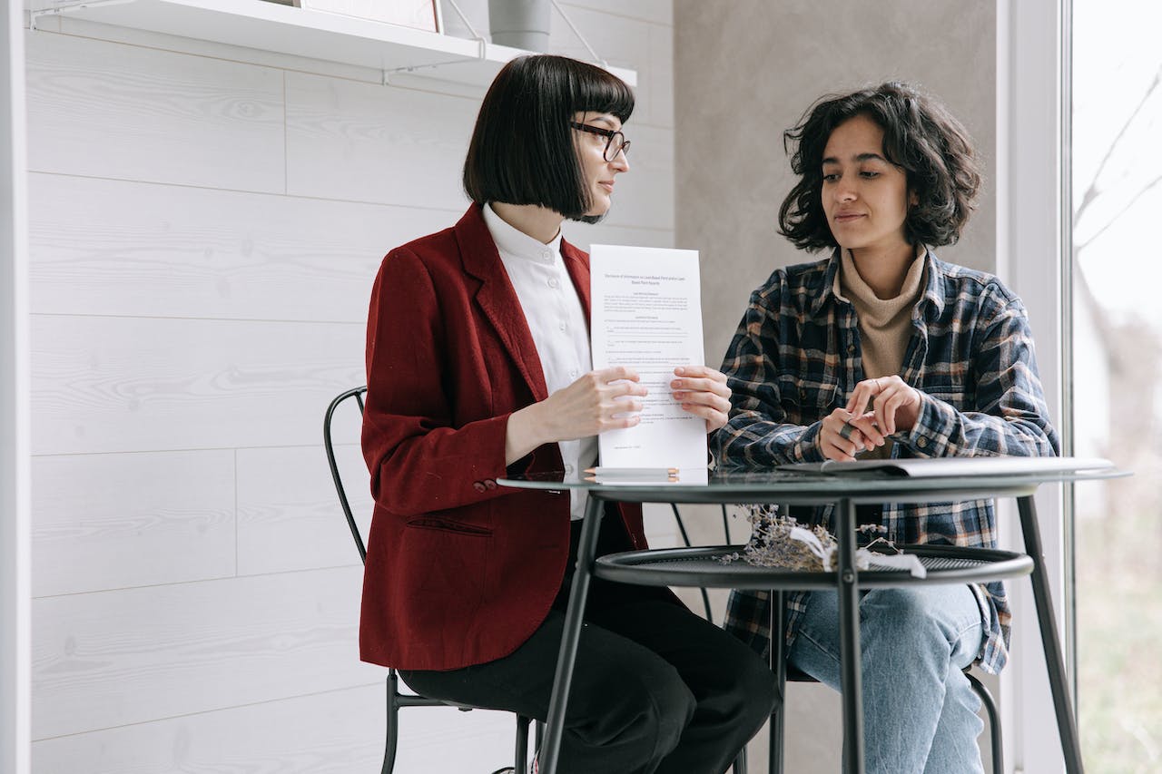 Woman in Red Blazer Holding Documents Beside Woman in Blue Button Up Plaid Shirt